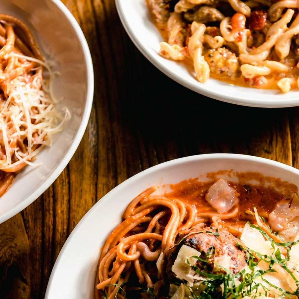 Three plates of saucy pasta dishes, including grated cheese, meatballs, herbs, and vegetables, arranged on a rustic wooden table.