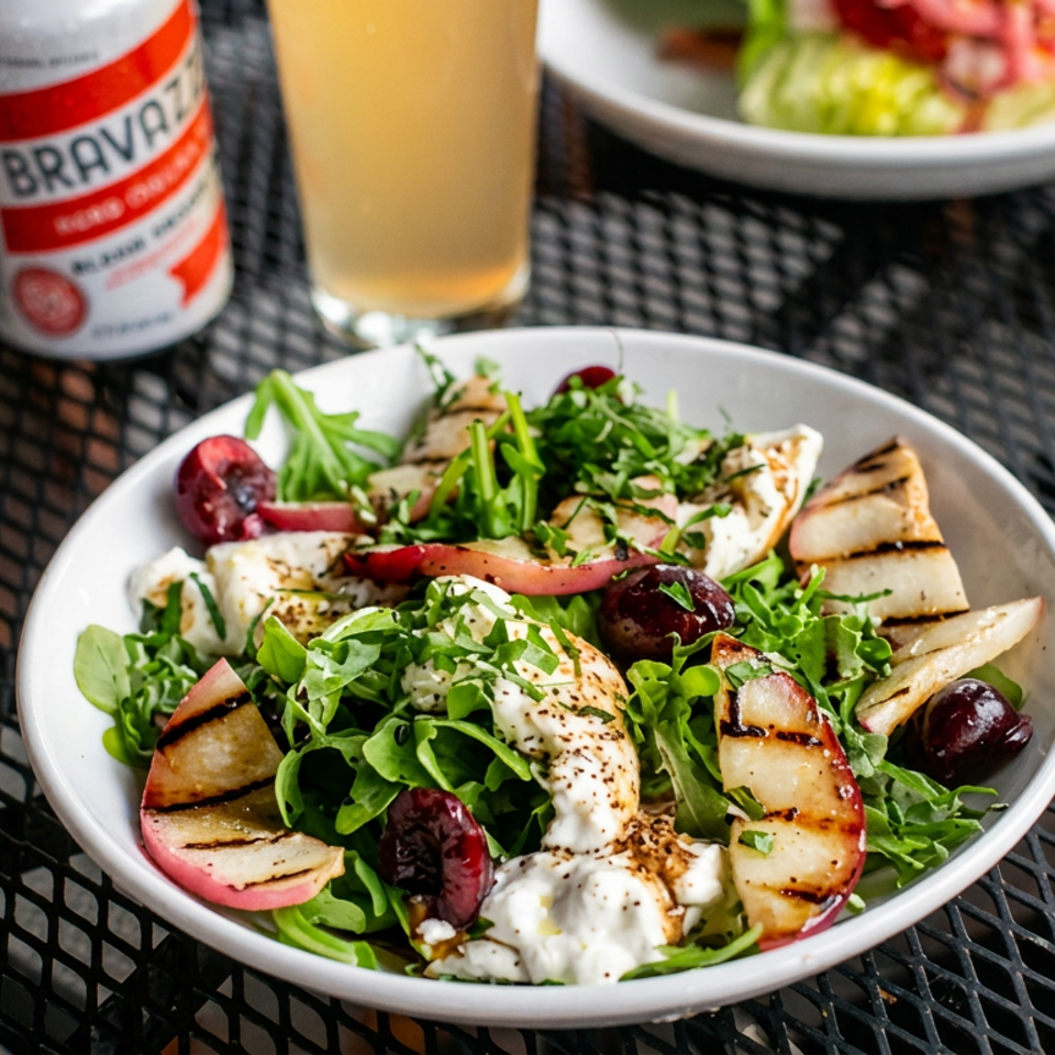 Mediterranean salad with grilled fruit, greens, olives, herbs, and creamy cheese served on an outdoor café table with a beer and lemonade.