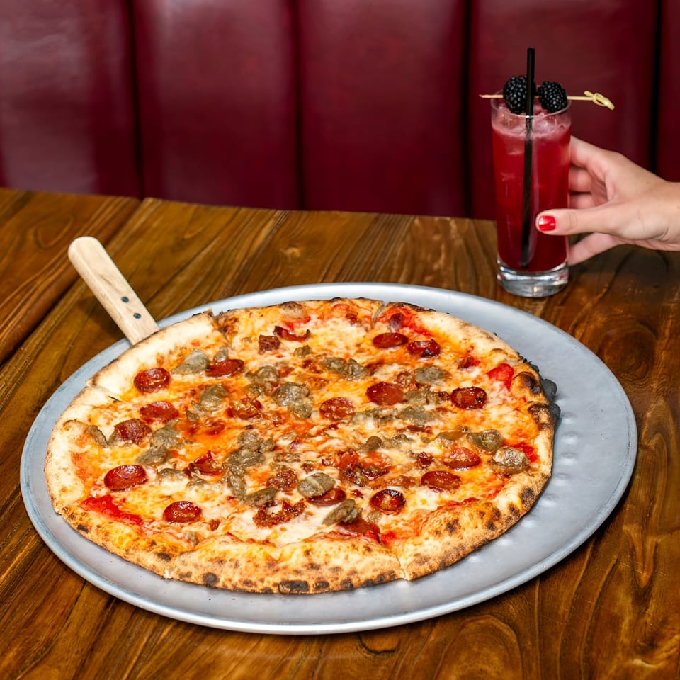Pepperoni and sausage pizza on a metal tray beside a red berry cocktail garnished with blackberries.