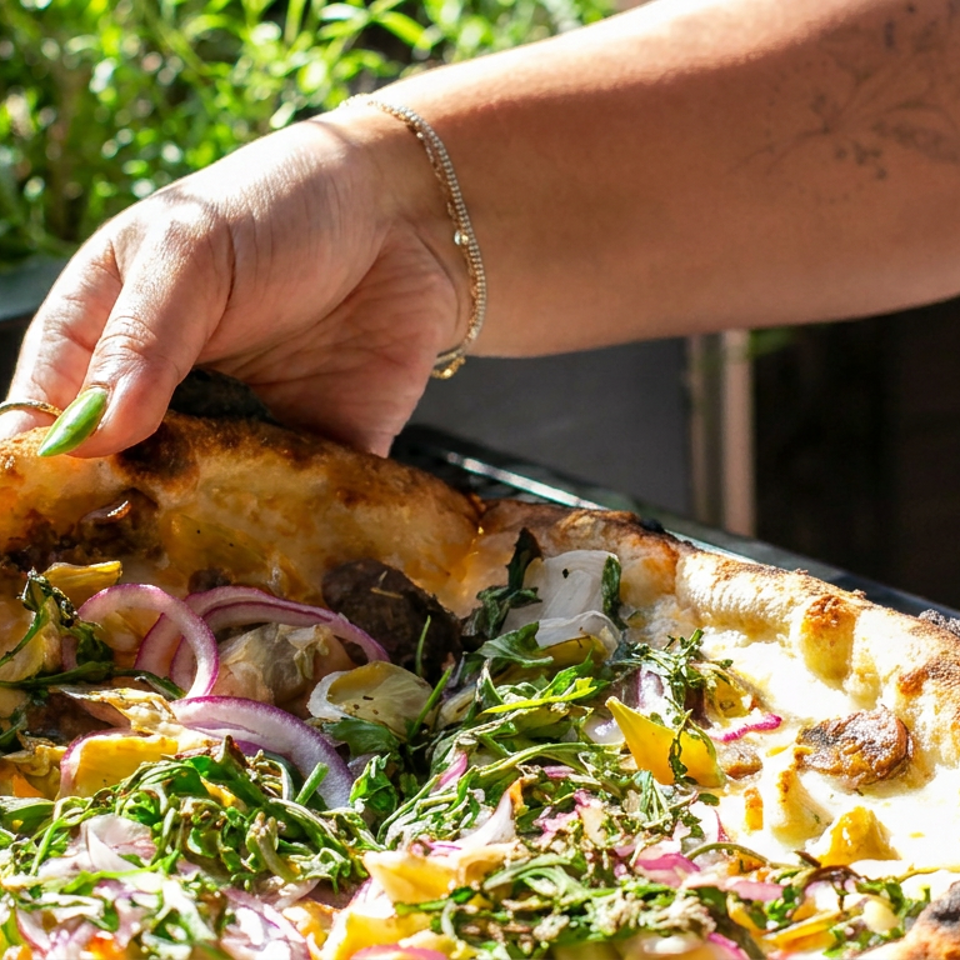 Hand lifts a golden flatbread topped with greens, red onion, herbs, and roasted vegetables outdoors.