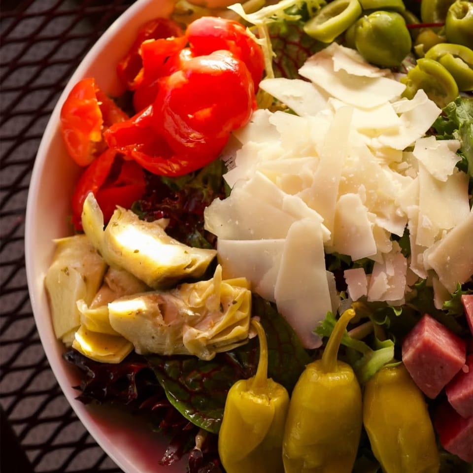 Close-up salad with leafy greens, tomatoes, olives, shaved Parmesan, artichoke hearts, pepperoncini, and diced meat.